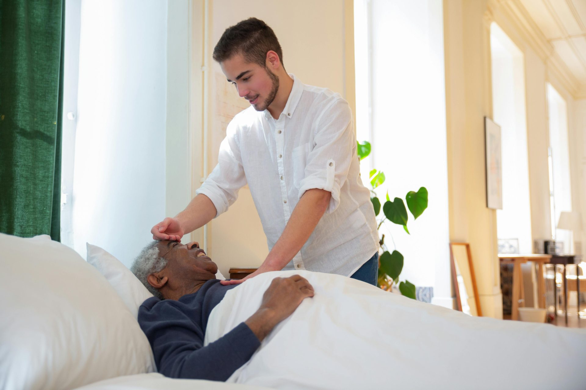 Ein junger Mann stützt einen älteren Mann im Bett und bietet ihm in einem hellen Zimmer Pflege an.